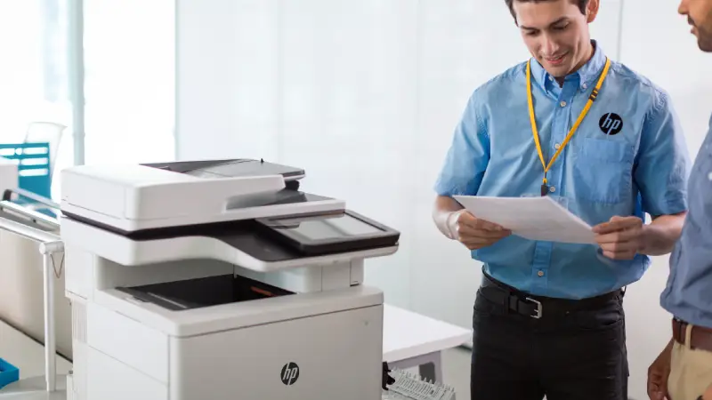 A person in a blue shirt and ID badge stands by a printer, reviewing documents in a bright office with modern furniture